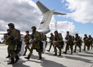 US military officials in uniform observing a military exercise with Belarusian or Russian counterparts, possibly shaking hands, with military vehicles or a map in the background. The atmosphere suggests a formal but tense interaction.
