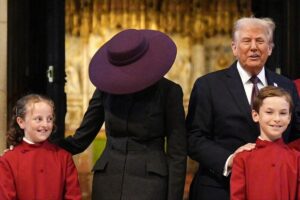 U.S. First Lady Melania Trump wearing a large, wide-brimmed hat at a state ceremony, with a subtle smile and a formal suit. The hat partially obscures her face.