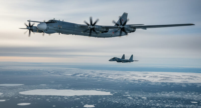 A wide-angle view of a Russian Tu-95MS strategic bomber flying over a vast, partly cloudy, icy sea, with a fighter jet faintly visible in the distance. The bomber has four turboprop engines and a distinctive large tail fin.