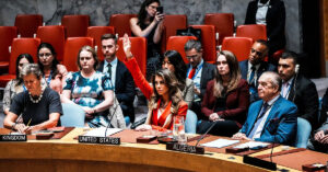 The United Nations Security Council meeting room with the flag of the United States prominently displayed in the foreground and a somber, concerned-looking group of diplomats representing various nations in the background. A red "VETO" stamp is superimposed on a draft resolution document on a table.