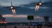 Two F-16 fighter jets rapidly taking off from an airfield at dusk, with afterburners glowing, as military personnel run on the tarmac. A radar screen and a control tower are visible, and a distant explosion or fire illuminates the horizon.