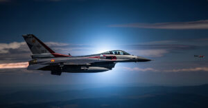 A dramatic photograph of a NATO F-16 fighter jet flying at night, with the silhouette of a drone faintly visible in the distance.