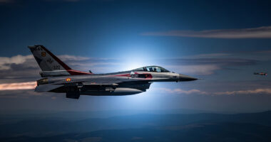 A dramatic photograph of a NATO F-16 fighter jet flying at night, with the silhouette of a drone faintly visible in the distance.