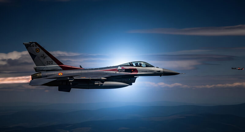 A dramatic photograph of a NATO F-16 fighter jet flying at night, with the silhouette of a drone faintly visible in the distance.