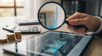 A person's hand with a magnifying glass examining a financial chart on a tablet, with a few coins and a calculator on a table. The image represents the careful scrutiny required for smart, low-risk investments.
