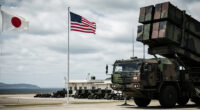 A powerful and serious image showing a modern missile launcher (like Typhon) on a military base in Japan, with Japanese and American flags visible. The background features a distant, subtly visible Chinese coastline, emphasizing the strategic deployment.