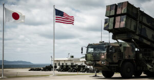 A powerful and serious image showing a modern missile launcher (like Typhon) on a military base in Japan, with Japanese and American flags visible. The background features a distant, subtly visible Chinese coastline, emphasizing the strategic deployment.
