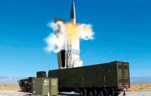 A powerful and serious image showing a modern missile launcher (like Typhon) on a military base in Japan, with Japanese and American flags visible. The background features a distant, subtly visible Chinese coastline, emphasizing the strategic deployment.